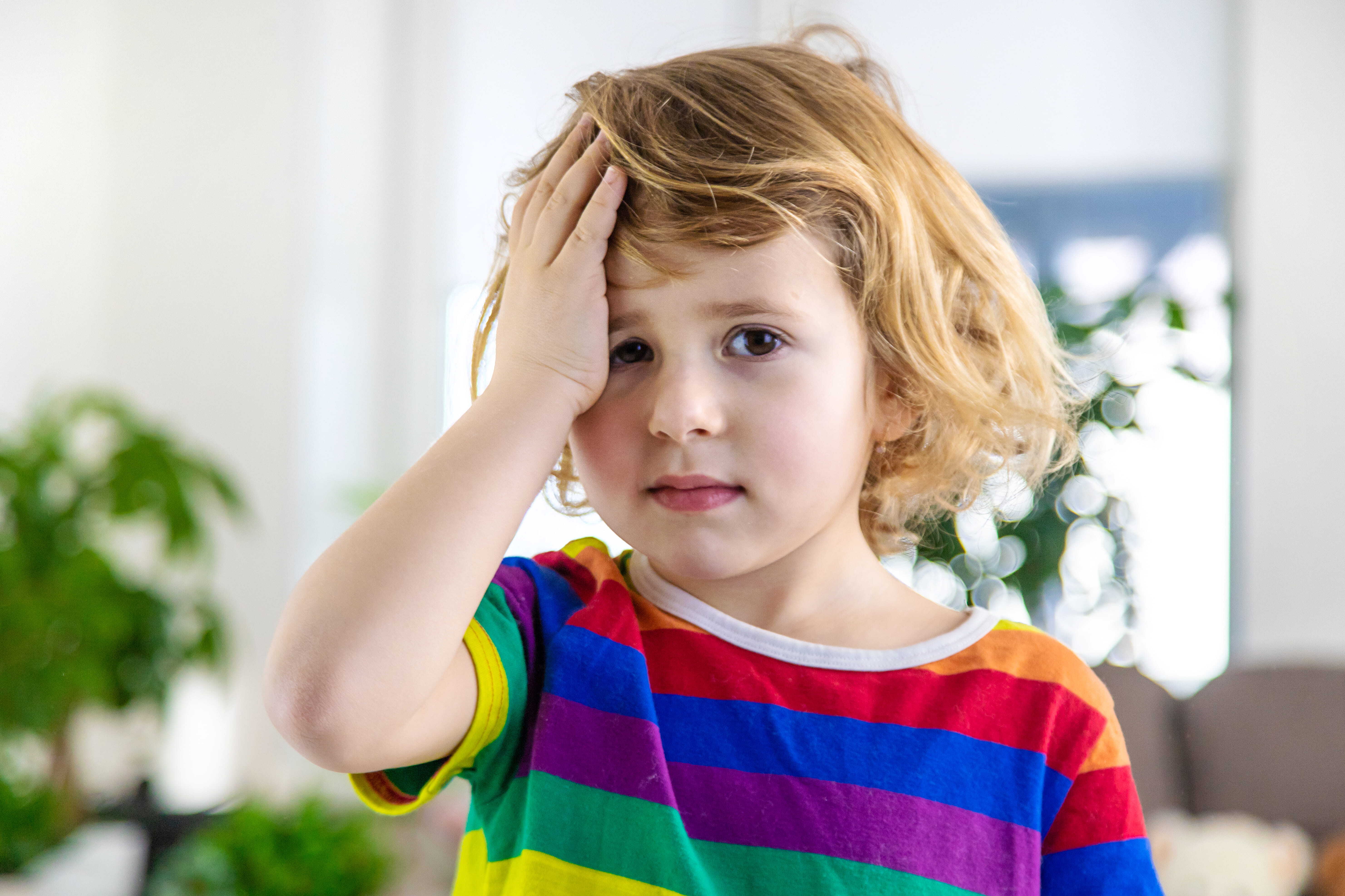A young child in a rainbow shirt with their hand to their face, looking at the camera, and appearing as though they have done something wrong.